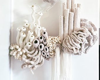 May include: A handmade textile art piece designed to be hung on a wall, representing an underwater scene. It features a crocheted fish, coral, and other marine life in white, beige, and grey tones. The artwork is mounted on a piece of driftwood.