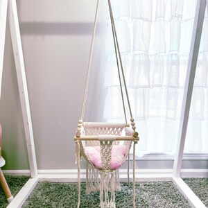 May include: A white macrame swing with a pink cushion hanging from a white wooden frame. The swing is in a room with a white rug and a white curtain.