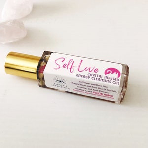 May include: A clear glass bottle with a gold cap and a pink label that reads "Self Love Crystal Infused Energy Cleansing Oil". The label also includes the text "Safflower and Rose Oils, Charged Clear and Rose Quartz Crystals, Jasmine, and Rose Flowers, Vitamin D, and GOOD VIBES".