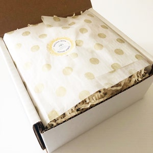 May include: A white box with a brown cardboard lid is open. The box is filled with brown packing paper and a white tissue paper with gold polka dots. The tissue paper has a sticker that says "Packed with Good Vibes" with a sun and a small leaf design.