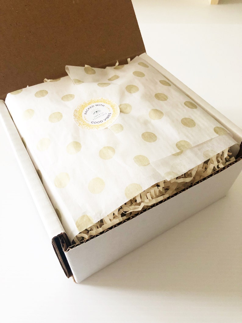 May include: A white box with a brown cardboard lid is open. The box is filled with brown packing paper and a white tissue paper with gold polka dots. The tissue paper has a sticker that says "Packed with Good Vibes" with a sun and a small leaf design.