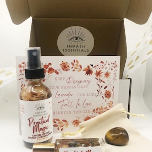 May include: A box with a white card that says "KEEP Rosemary YOUR GARDEN GATE Lavender FOR LUCK Fall In Love HENEVER YOU CAN" with a floral design. The box contains a clear bottle with a gold lid and a label that says "EMPATH ESSENTIALS Practical Magic CRYSTAL INFUSED ENERGY CLEANSING SPRAY 2 OZ / 60 ML", a smaller clear bottle with a gold lid and a label that says "Practical Magic CRYSTAL INFUSED ENERGY CLEANSING OIL", and a brown tiger's eye stone in a small white pouch.