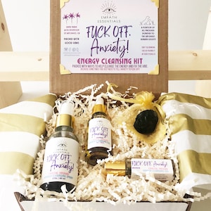 May include: A white box filled with gold and white shredded paper. The box contains three glass bottles with gold lids and labels that say "Fuck Off Anxiety!"  The bottles contain crystal infused energy cleansing spray and oil. There is also a small black stone in a gold mesh bag.