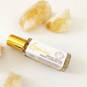 May include: A glass roller bottle with a gold cap and a white label that says "Gemini Crystal Infused Energy Cleansing Oil". The label also includes the text "Safflower Oil and Citrus Essential Oils, Align with Gemstones and Crystals, Grounding, Harmony and Angelic Flowers". The bottle is sitting on a white surface with several yellow crystals.