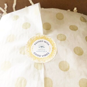 May include: A white paper bag with gold polka dots and a sticker that says "Packed with Empath Essentials Good Vibes". The sticker has a gold glitter background and a blue eye symbol.