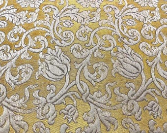 May include: Close-up of a gold-colored fabric with a repeating floral pattern in off-white. The design features stylized flowers, leaves, and swirling vines, creating a textured, ornate look. The fabric has a subtle sheen.