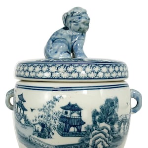 May include: A blue and white ceramic jar with a lid featuring a seated dog figurine. The jar is decorated with a landscape scene in blue, including pagodas, trees, and a bridge. It has two small handles on the sides.