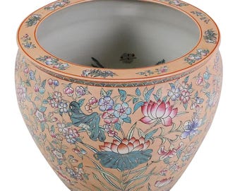 May include: A peach-colored ceramic planter with a wide, round shape. The exterior is adorned with a detailed floral pattern featuring pink, blue, and green flowers and leaves. The rim has a decorative border, and the interior is a light gray color.