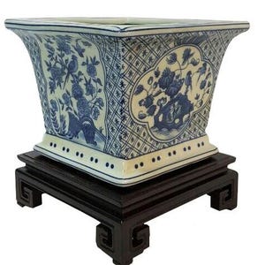 May include: A square, blue and white porcelain planter with floral and bird designs, set on a dark wooden stand. The planter has a flared top and is decorated with intricate patterns and images.