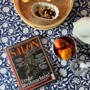 Könnte beinhalten: Eine festliche Szene mit einem Magazin, Obst und Snacks auf einer blauen Tischdecke mit Blumenmuster. Ein geflochtener Tablett hält Kerzen und eine Schale mit Nüssen. Das Magazin zeigt "Frohe Weihnachten!"