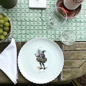 Könnte beinhalten: Ein weißer Porzellanteller mit einem gewellten Rand, der eine Vintage-Illustration eines Mannes zeigt, der Wein verkauft. Die Illustration ist farbig und zeigt den Mann, der einen Korb mit Weingläsern auf dem Rücken trägt. Der Teller trägt den Text "Since 1872" und "Marchand de Vin".