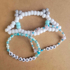 May include: Three beaded bracelets with white, blue, and gray beads. The bracelets spell out "NED BAYOU" and "NED".