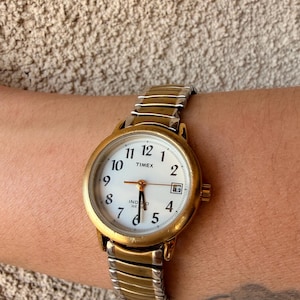 Puede incluir: Un reloj de pulsera Timex con caja redonda de tono dorado y esfera blanca con números arábigos negros. El reloj cuenta con una ventana de fecha en la posición de las tres y una correa extensible de dos tonos, dorada y plateada, de metal.