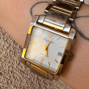 May include: A rectangular wristwatch with a two-tone silver and gold metal link band. The watch face is white with gold-colored hour markers, gold hands, and the text Precision Quartz by GRUEN. It features a date window at the three o'clock position.