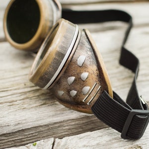 Op de afbeelding: Steampunk-bril met een bruin leren frame, zilveren accenten en een verstelbare zwarte band. De bril heeft een verweerde look en is gemaakt van metaal en leer.