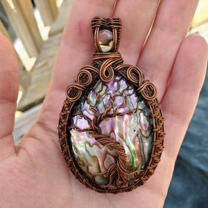 May include: A handcrafted copper wire pendant featuring an abalone shell with a tree of life design. The pendant has a detailed wire-wrapped frame and bail, showcasing iridescent colours of blue, green, and purple.