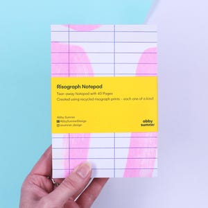 May include: A white notepad with a grid pattern and pink abstract shapes, held by a hand. The notepad has a yellow label with the text "Risograph Notepad" and details about the product. The notepad is tear-away with 40 pages.