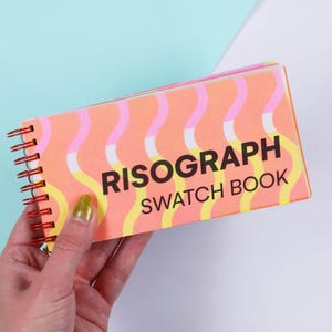 May include: A rectangular spiral-bound sketchbook with a peach-coloured cover featuring a repeating pattern of vertical wavy lines in pink, white, and yellow. The text RISOGRAPH SWATCH BOOK is printed in bold black capital letters on the front cover.