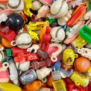 Puede incluir: Una colección de pequeños amuletos y baratijas de plástico de colores. El surtido incluye pelotas de béisbol, perritos calientes, manzanas, barcos y otros artículos de novedad en miniatura en varios colores como rojo, amarillo y blanco. Estos amuletos son probablemente para manualidades o fabricación de joyas.