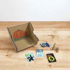 May include: A cardboard box decorated with a pumpkin painting, with miniature Halloween gift bags. The bags display a black cat, a ghost, and a witch, with the words "Happy Halloween!" on them. A small bicycle and other miniature items are also present.