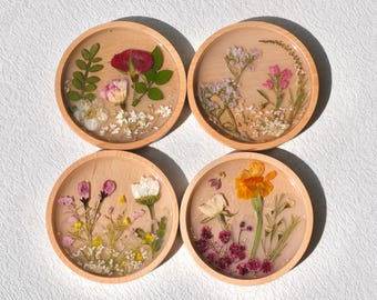 May include: Four round wooden coasters with pressed flowers and leaves encased in clear resin. The coasters feature a variety of flowers in shades of pink, white, yellow, and red.