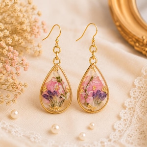 May include: A pair of teardrop-shaped earrings featuring gold-toned metal frames. Inside the clear resin, there are pressed pink and purple flowers accented with small flecks of gold leaf. The earrings hang from simple gold-toned metal hooks.