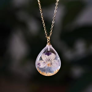 May include: A gold chain necklace with a clear teardrop pendant containing a pressed white flower with purple accents.