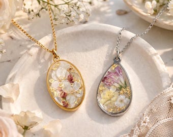 May include: Two necklaces with resin pendants. One is an oval gold-tone pendant containing pressed white and pink flowers with gold flakes on a gold chain. The other is a teardrop silver-tone pendant with purple, yellow, and white flowers.
