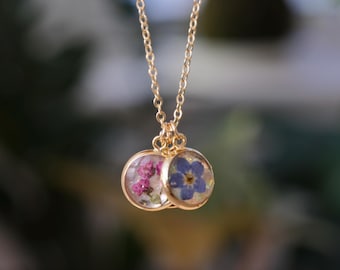 May include: A gold-colored necklace with two round pendants. One pendant contains pink flowers, and the other features a blue flower. The chain and pendants are delicate, creating an elegant floral jewelry piece.