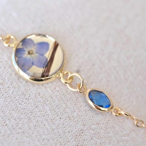Puede incluir: Pulsera dorada con una flor azul prensada en un engaste redondo y una gema azul. La cadena es fina y elegante. La pulsera es una joya. Diseño sencillo y delicado, adecuado para el uso diario.