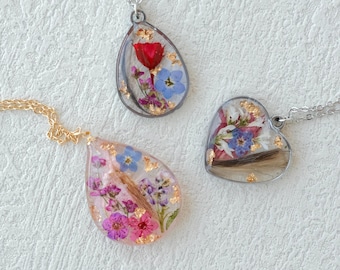 May include: Three resin pendant necklaces with dried flowers and gold flakes. One is teardrop-shaped with pink and purple flowers, another is a teardrop with a red flower, and the last is heart-shaped.
