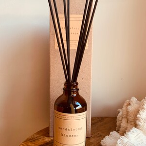 May include: A brown glass bottle with a label that reads "sandalwood blossom reed diffuser". The bottle is filled with a clear liquid and has black diffuser sticks sticking out of the top. The bottle is sitting on a wooden surface.