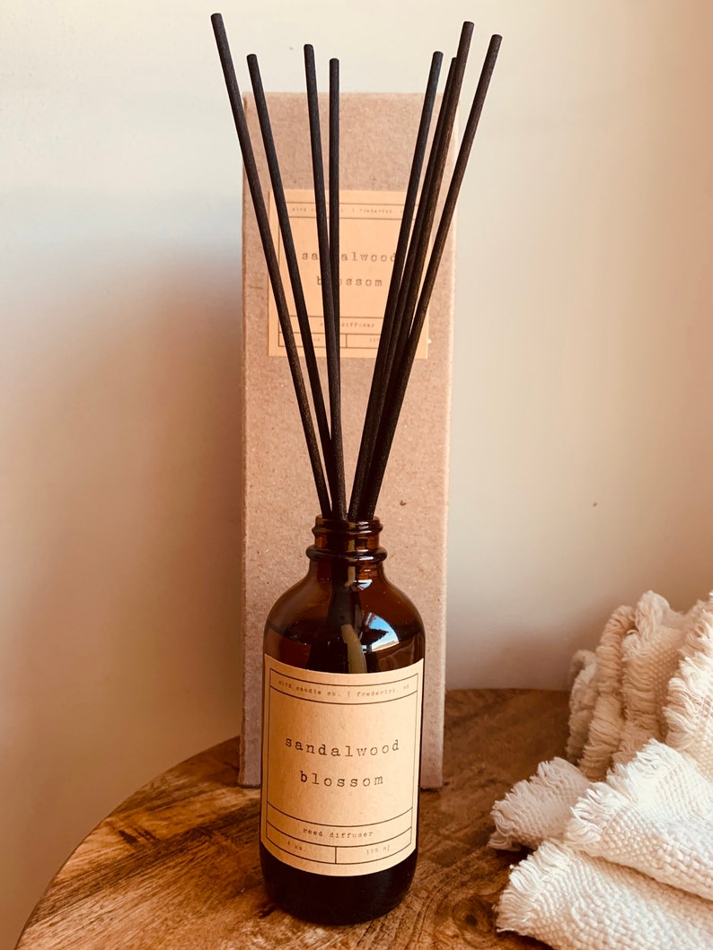 May include: A brown glass bottle with a label that reads "sandalwood blossom reed diffuser". The bottle is filled with a clear liquid and has black diffuser sticks sticking out of the top. The bottle is sitting on a wooden surface.