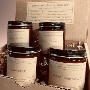 May include: A botanical candle sampler set in a cardboard box. The set includes four glass jars with black lids, each labeled with a different scent: honeysuckle, lilac blooms, rosewater, and sweet magnolia. The box also contains a printed card listing the scents.
