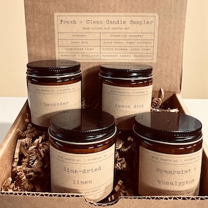 May include: A candle sampler set in a cardboard box, featuring four glass jars with black lids. Each jar has a label with a scent name: lavender, lemon drop, line-dried linen, and spearmint + eucalyptus. The box includes a scent list.