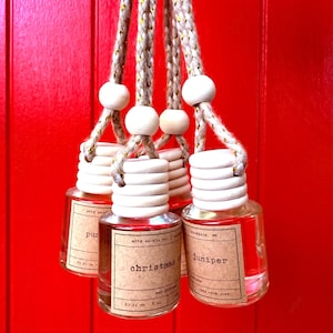 May include: Several small, clear glass bottles with wooden tops and hanging cords. Each bottle has a label with text, including "christmas tree" and "Juniper". The bottles are filled with a reddish-brown liquid and hang against a bright red background.