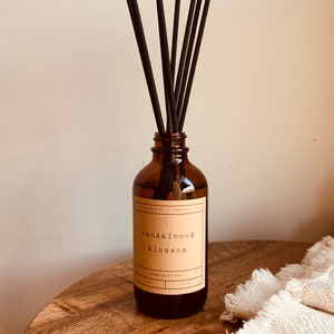 May include: A brown glass bottle with a brown label that reads "sandalwood blossom reed diffuser". The bottle is filled with a clear liquid and has black reeds sticking out of the top.