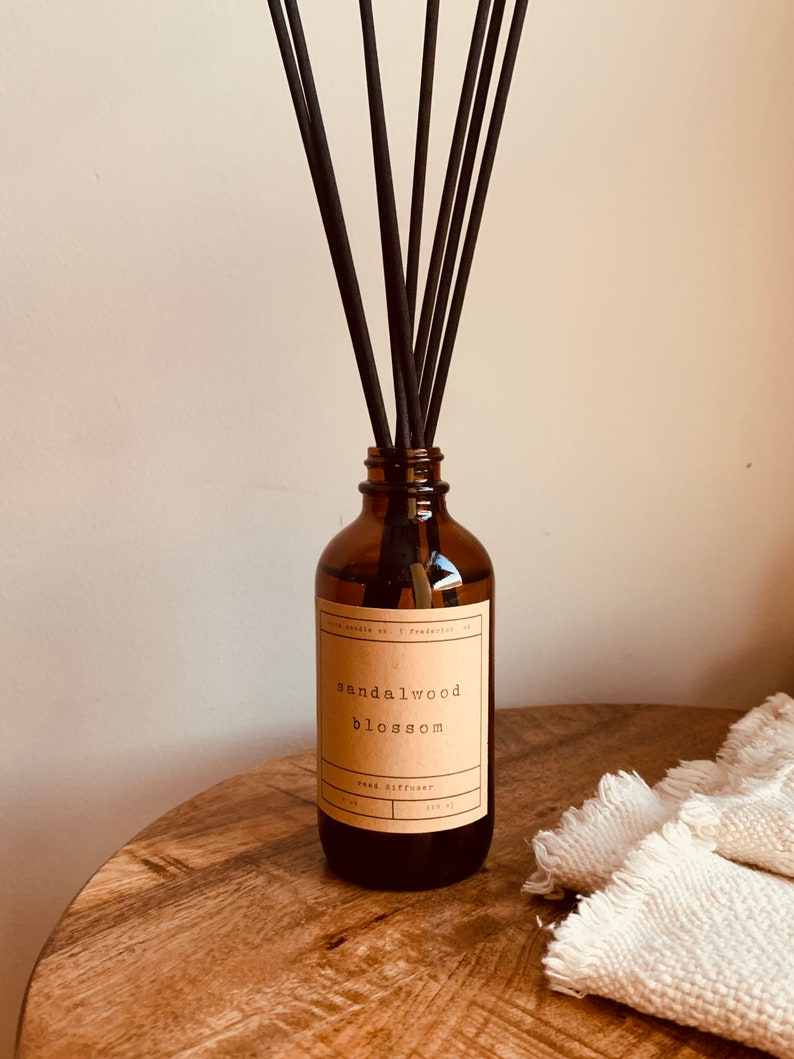 May include: A brown glass bottle with a brown label that reads "sandalwood blossom reed diffuser". The bottle is filled with a clear liquid and has black reeds sticking out of the top.