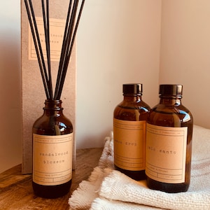 May include: Three brown glass bottles with black labels and brown reed diffusers. The labels read "sandalwood blossom", "lemon drop", and "palo santo". The bottles are sitting on a white fabric surface.