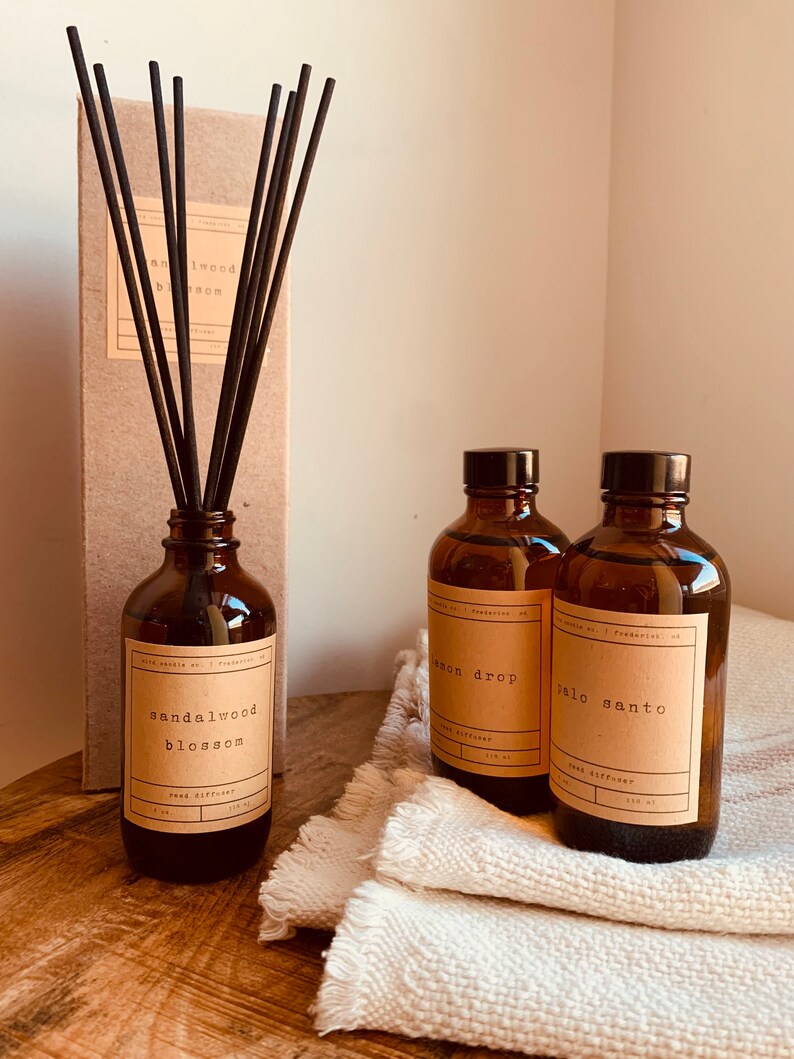 May include: Three brown glass bottles with black labels and brown reed diffusers. The labels read "sandalwood blossom", "lemon drop", and "palo santo". The bottles are sitting on a white fabric surface.