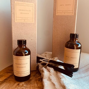 May include: Two brown cardboard boxes with white labels that say "sandalwood blossom" and "palo santo" reed diffusers. The boxes are next to two brown glass bottles with the same labels. The bottles are sitting on a wooden surface with a white towel and black reed diffuser sticks.