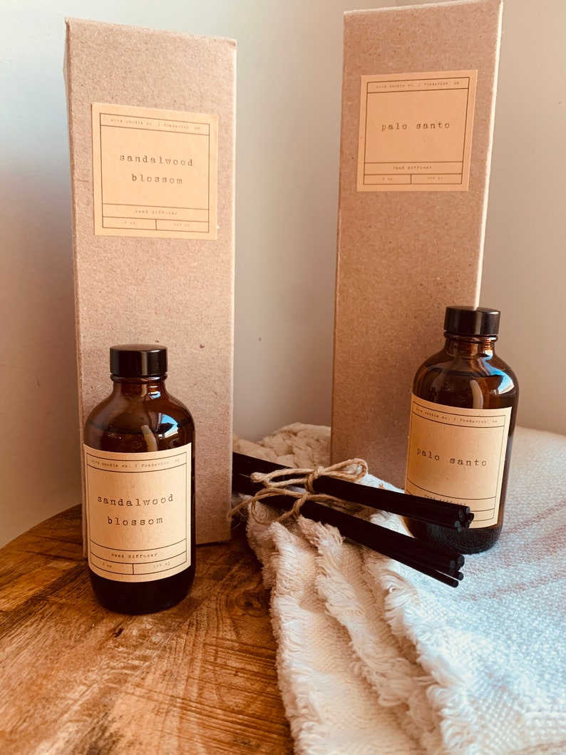 May include: Two brown cardboard boxes with white labels that say "sandalwood blossom" and "palo santo" reed diffusers. The boxes are next to two brown glass bottles with the same labels. The bottles are sitting on a wooden surface with a white towel and black reed diffuser sticks.