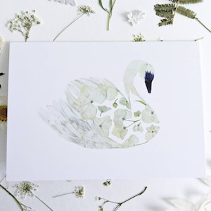 May include: A white greeting card featuring a swan illustration made of pressed flowers. The swan has a blue beak and is surrounded by dried flowers and leaves. The card is on a white surface.