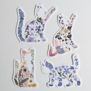 Puede incluir: Cuatro pegatinas con forma de gato hechas de flores prensadas. Los gatos est&aacute;n en varias poses y est&aacute;n decorados con flores moradas, azules, rosas y blancas.