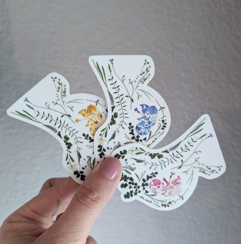 May include: Three white stickers featuring pressed flowers in the shape of birds. The flowers are yellow, blue and pink.
