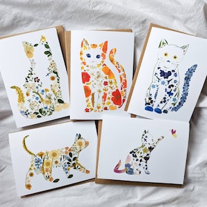 May include: Five greeting cards featuring cat silhouettes filled with colorful pressed flowers. The cats are in various poses, with a butterfly on one card. The cards have a white background and brown backing.