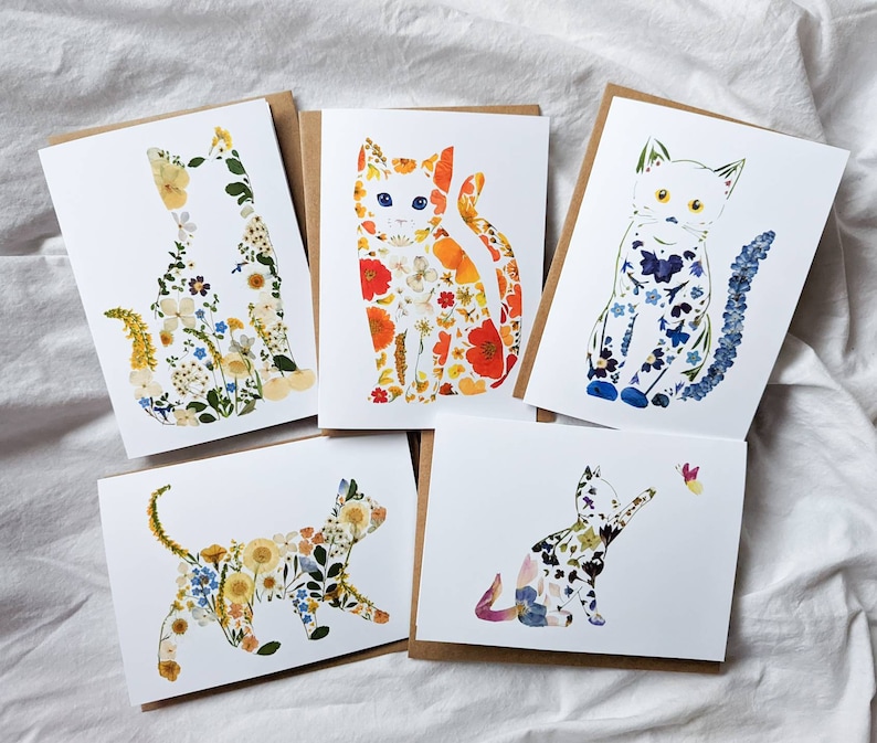 May include: Five greeting cards featuring cat silhouettes filled with colorful pressed flowers. The cats are in various poses, with a butterfly on one card. The cards have a white background and brown backing.
