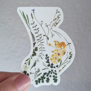 May include: A white sticker featuring a silhouette of a pregnant belly filled with pressed flowers and leaves. The flowers are yellow, green, and blue.