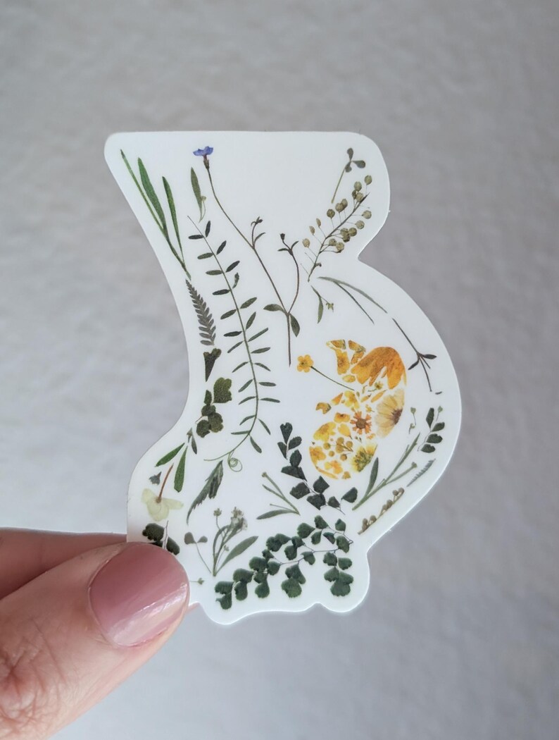 May include: A white sticker featuring a silhouette of a pregnant belly filled with pressed flowers and leaves. The flowers are yellow, green, and blue.