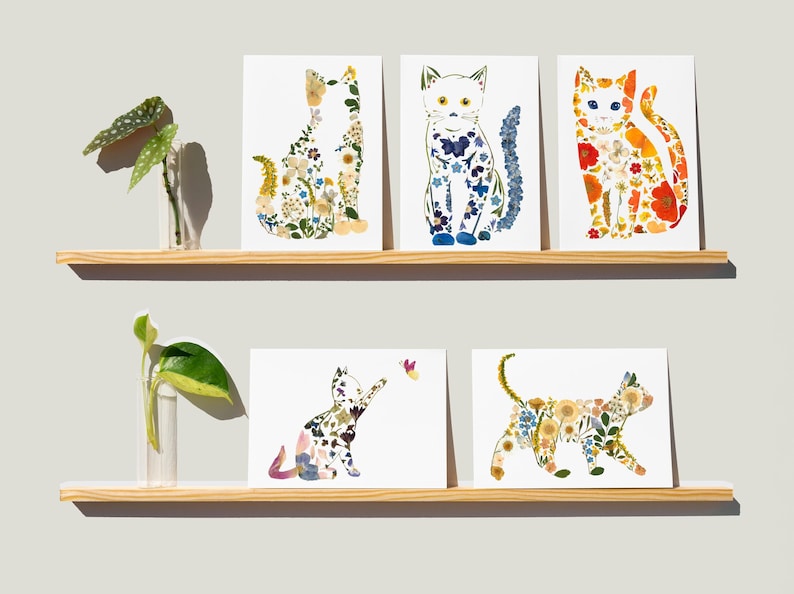 May include: Six white art prints featuring cat silhouettes composed of colorful pressed flowers. The prints are displayed on two wooden shelves, with small glass vases holding green plants on each shelf. The cats are in various poses.